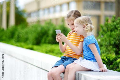 Obraz na płótnie Two cute little sisters playing outdoor mobile game on their smart phones