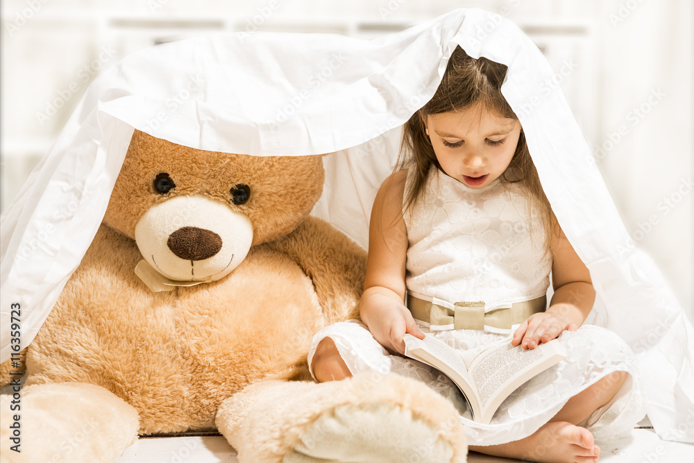 Beautiful little girl reading to her teddy bear toy friend under the blanket