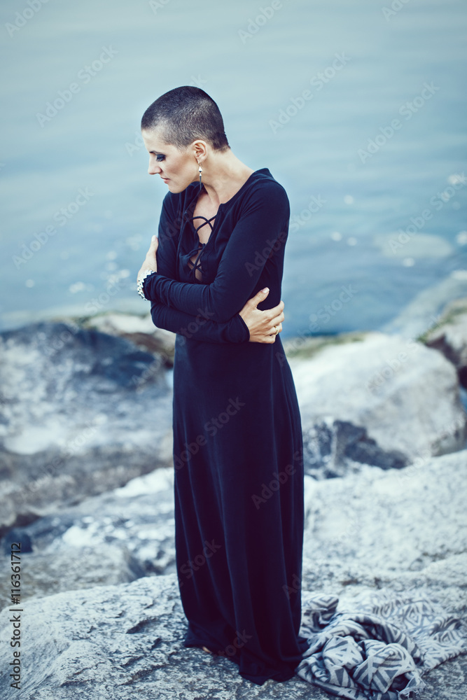 Portrait of sad beautiful Caucasian white young bald girl woman with shaved hair head standing by water lake looking away, mystic dark mood, toned with Instagram filters in blue green color Portrait of sad beautiful Caucasian white young bald girl woman with shaved hair head standing by water lake looking away, mystic dark mood, toned with Instagram filters in blue green color