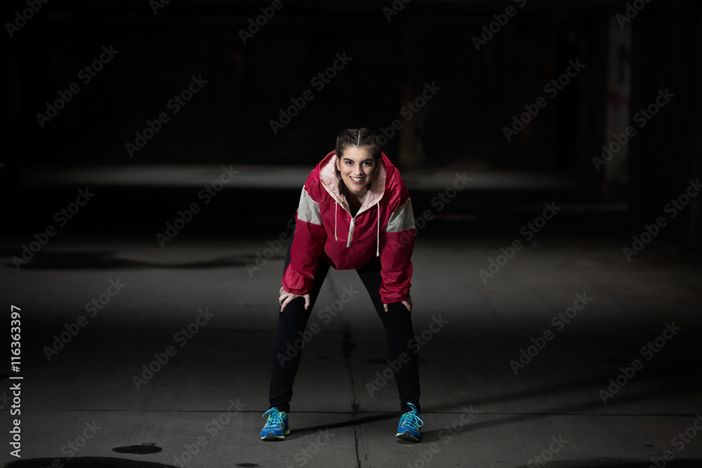 Portrait of a teenage girl resting after exercising in an urban environment. She is wearing magenta sweatshirt as a contrast to dark gray concrete underground garage.