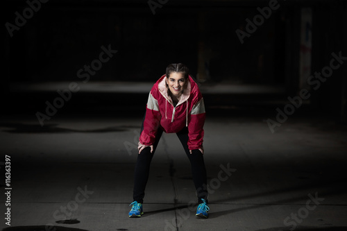 Portrait of a teenage girl resting after exercising in an urban environment. She is wearing magenta sweatshirt as a contrast to dark gray concrete underground garage.