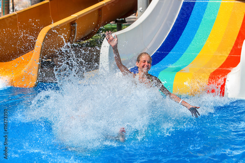 Happy kid on water slide at water park hands up. There are some water slides with flowing splash water in aqua park. Summer water park holiday. Outdoor. © Gennadiy Poznyakov