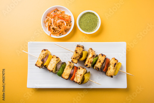 Paneer Tikka Kabab - Tandoori Indian cheese skewers, malai paneer tikka / malai paneer kabab, chilli paneer served in white plate with barbecue stick and colourful capsicum and onion, with green sauce photo