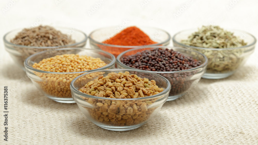 Different types of Indian spices in glass bowl, focus on Fenugreek seeds on jute mat background. Front view.