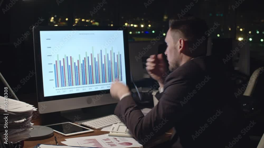 Medium shot of exhausted businessman analyzing financial statistics on computer screen, taking off his glasses, trying to concentrate and drinking coffee in dark office late at night Medium shot of exhausted businessman analyzing financial statistics on computer screen, taking off his glasses, trying to concentrate and drinking coffee in dark office late at night