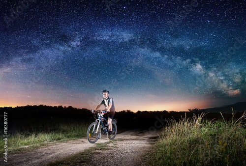 Sporty man riding bicycle in the night under Milky way far away from city. Starry Sky shines above the rider