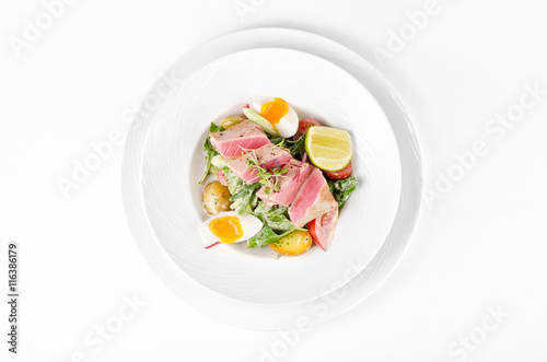 Salad with tuna, lettuce, tomatoes, eggs and potatoes on a plate on a white background, top view