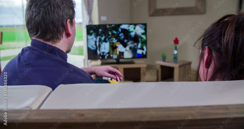 A man is watching tv and then he is looking into the camera. His wife continues watching some action movie. Close-up shot.
A man is watching tv and then he is looking into the camera. His wife continues watching some action movie. Close-up shot.