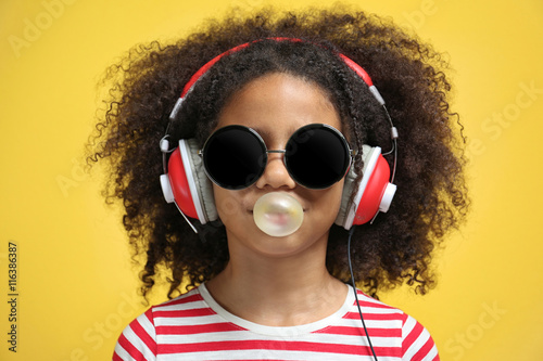 Obraz na plátně Afro-American little girl with headphones and sunglasses chewing gum on yellow b