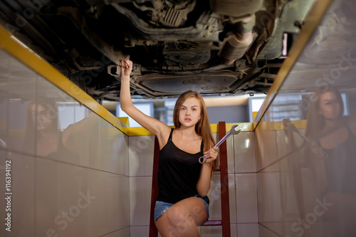 Young sexy brunette mechanic in checked shirt in car repair pit.