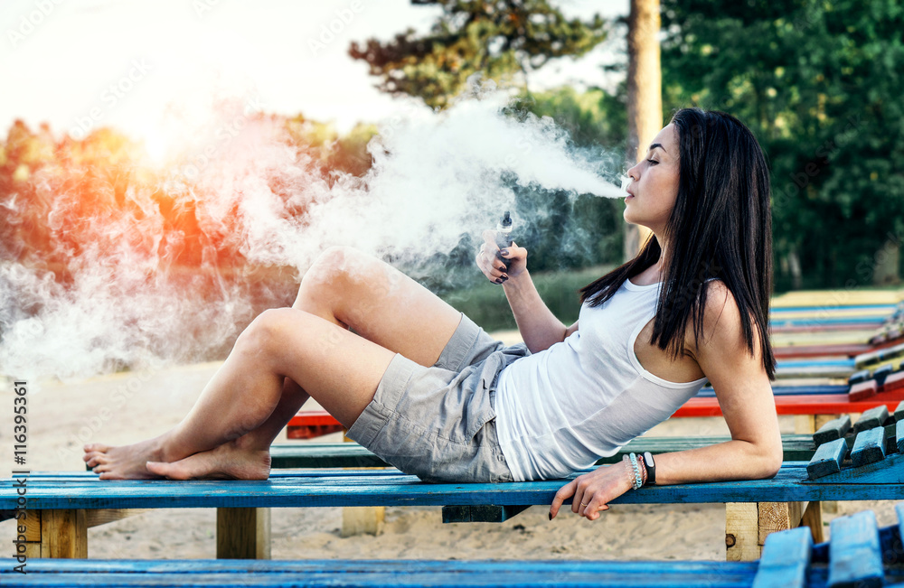 Naklejka premium Long hair brunette girl smoking electronic cigarette on the beac