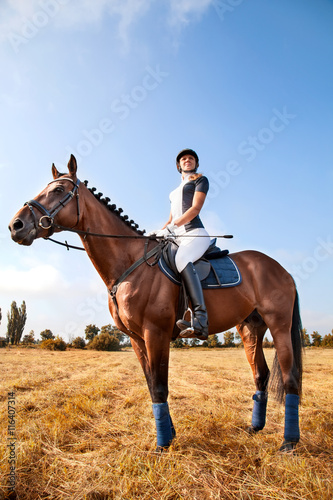 Cheerful beautiful young girl jockey in uniform sitting on a horse against blue sky and yellow field,looking forward and smilling on a sunny day. Equestrian sport - dressage.