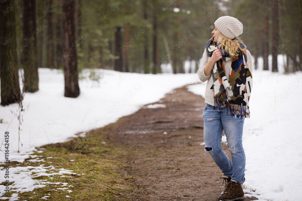 Young blond girl in blue jeans, warm sweater and bright scarf standing in a winter forest. Winter walks outdoors. Young blond girl in blue jeans, warm sweater and bright scarf standing in a winter forest. Winter walks outdoors.