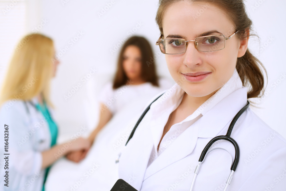 Fototapeta premium Young beautiful female doctor smiling on the background with patient and his physician in hospital. High level and quality medical service concept.