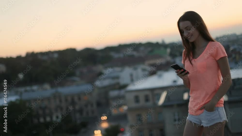 Moving view of brunette woman reading messages on smartphone at roof top, cityscape at sunset as background Moving view of brunette woman reading messages on smartphone at roof top, cityscape at sunset as background