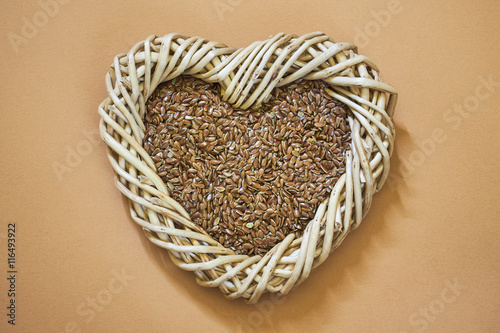 Flax seed in woven heart-shaped basket on light brown background. Copy space