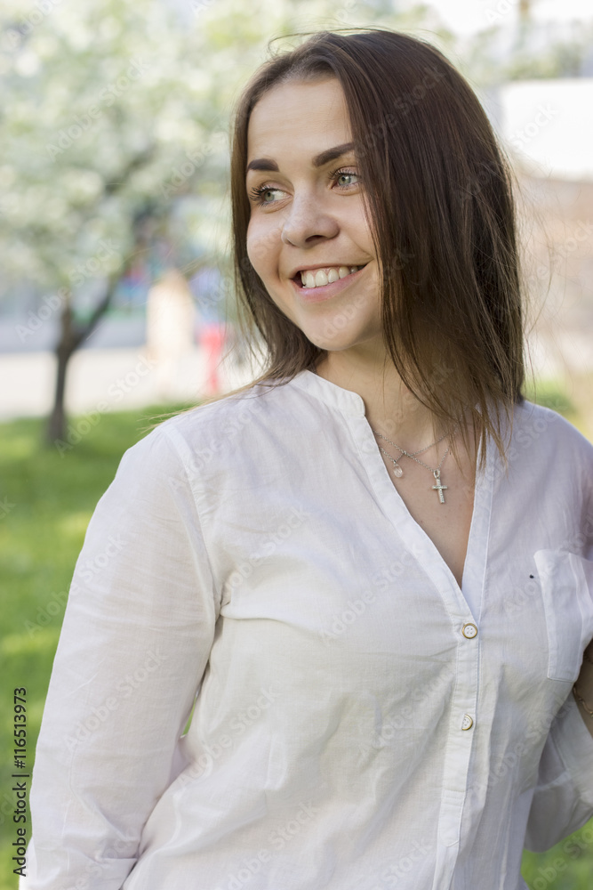 Young beautiful woman in a white shirt standing by a tree on the park looking sideways smiling Young beautiful woman in a white shirt standing by a tree on the park looking sideways smiling
