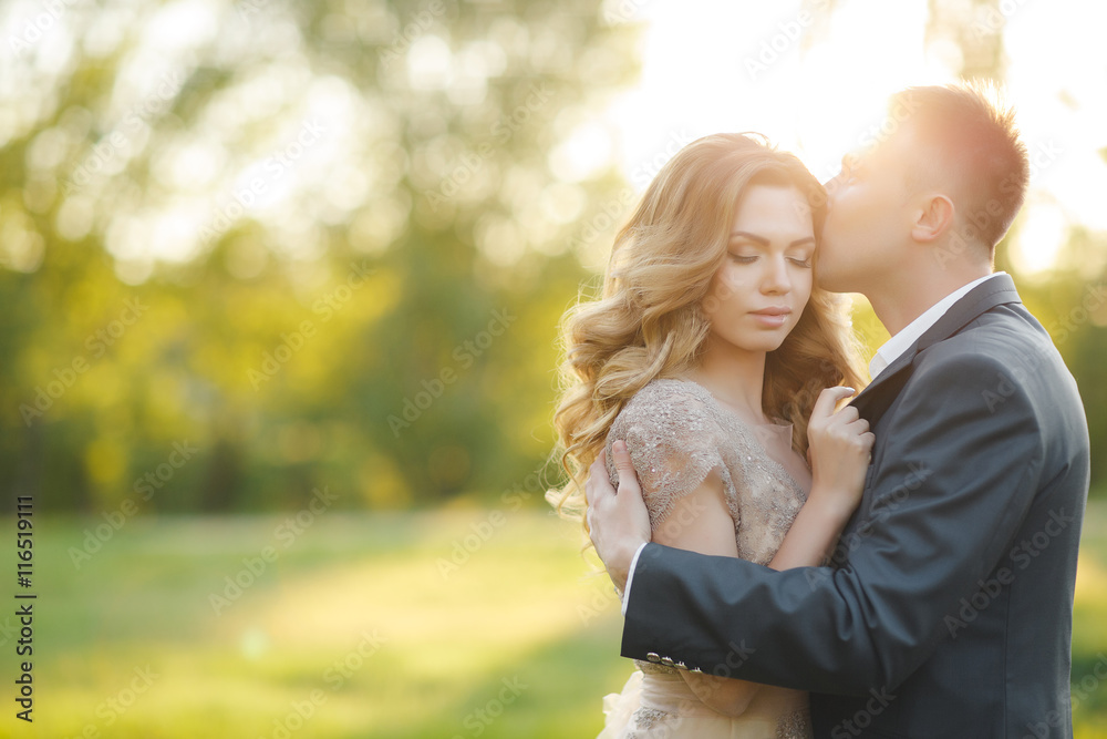 Fototapeta premium A young couple in love,bride and groom,blonde woman and man in wedding attire,standing arm in arm on a green summer meadow at sunset,romantic moments of the wedding