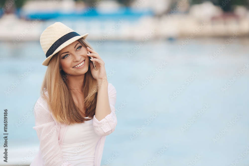 Girl in a beige hat,a beautiful smile,gray eyes,white straight teeth,long blonde straight hair,wearing a light shirt, talking on mobile phone sitting on the embankment near the blue ocean