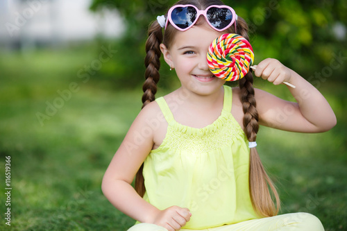 The girl with pigtails,wearing sunglasses in pink frame with glass in the form of hearts,dressed in a yellow t-shirt and pants sitting on green grass in summer Park with a large round candy on a stick