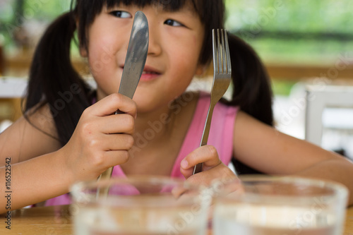 little asian girl waiting for dinner. Holding a knife and fork in the hand

