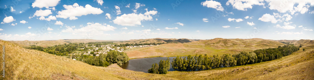 Summer panorama of the hilly terrain of the steppe lake, trees and village.