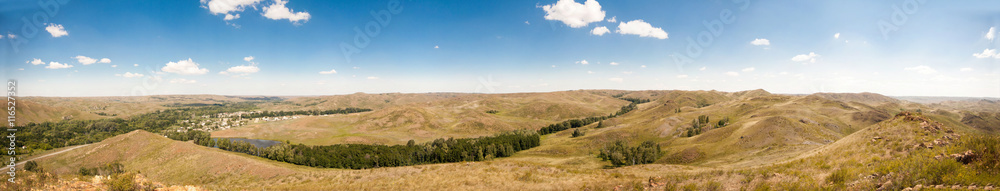 Summer panorama of the hilly terrain of the steppe lake, trees and village.