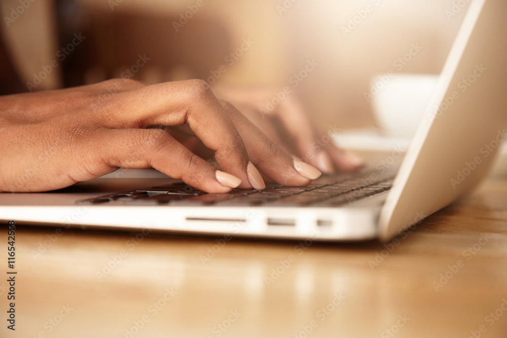 Cropped portrait of young female student learning on-line using generic laptop computer. Dark-skinned business woman sitting in front of open net-book, typing a message. Selective focus on fingers