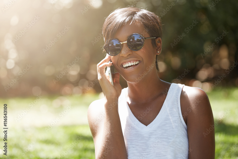 Close up portrait of dark skinned female laughing at a joke while having a phone conversation, enjoying summer sunny day in the city park, wearing stylish sunglasses, looking carefree and happy Close up portrait of dark skinned female laughing at a joke while having a phone conversation, enjoying summer sunny day in the city park, wearing stylish sunglasses, looking carefree and happy