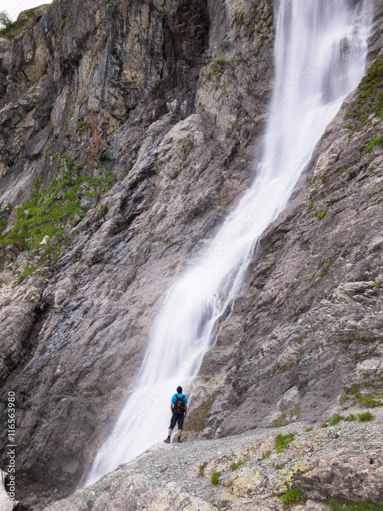 Adult male with a backpack standing lonely on the edge of the cliff and looking at the big waterfall