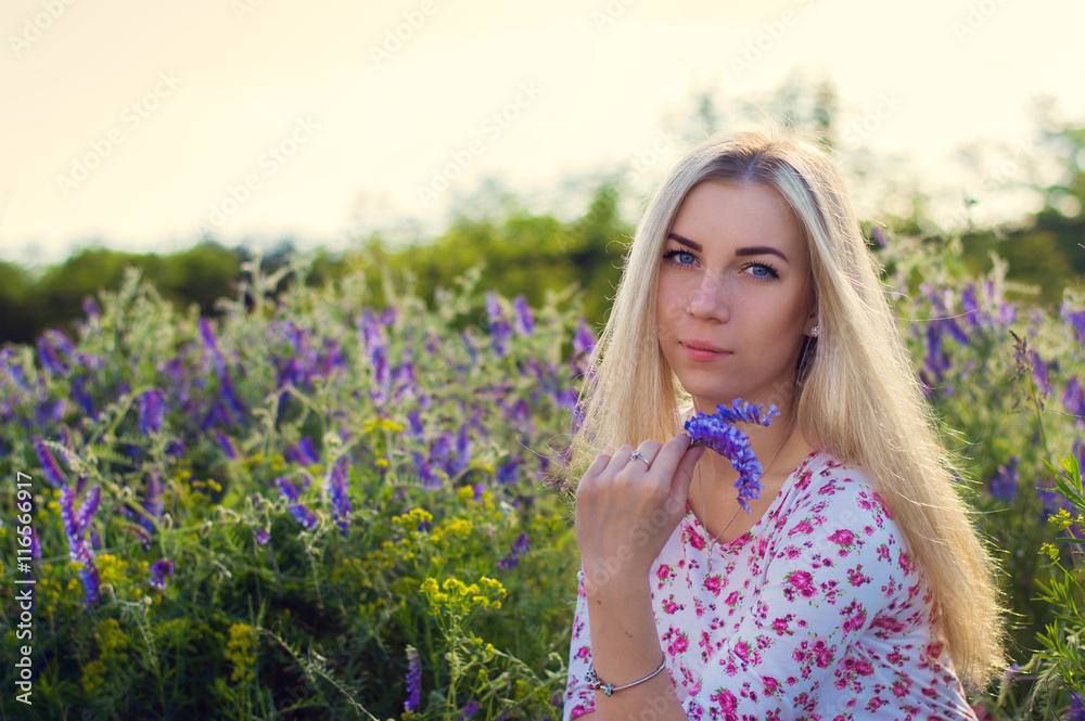 Blond girl with beautiful big blue eyes sitting in the field with purple flowers Blond girl with beautiful big blue eyes sitting in the field with purple flowers