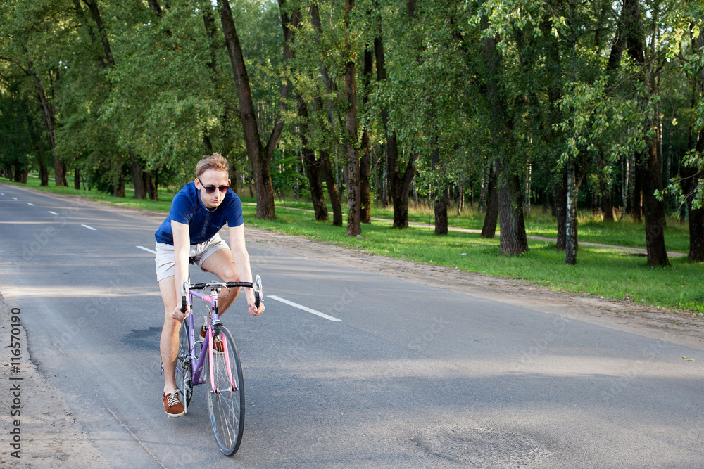 Young trendy man in sunglasses riding a bike on the asphalted road near the city park