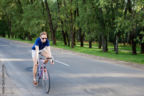 Young trendy man in sunglasses riding a bike on the asphalted road near the city park
