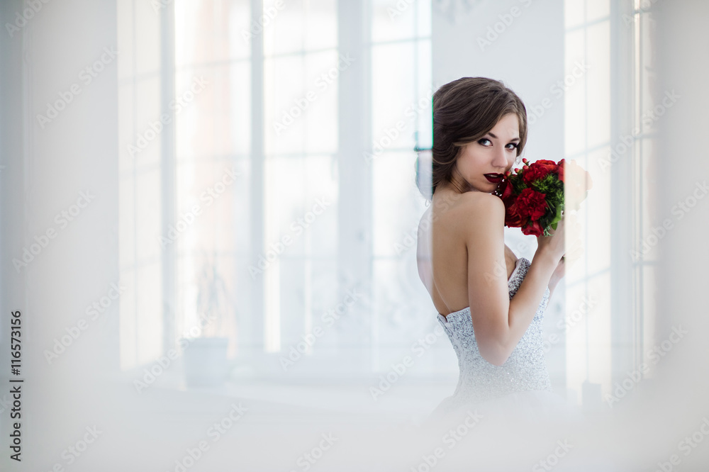 Glamorous young bride with flowers wearing wedding dress standing in front of doors indoors at luxury interior room Glamorous young bride with flowers wearing wedding dress standing in front of doors indoors at luxury interior room