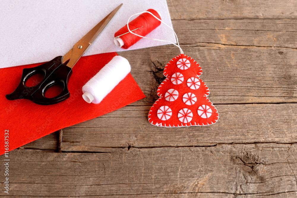 Red felt Christmas tree decor, scissors, red and white felt sheets, thread,  needle on wooden background with empty space for text. Home sewing crafts.  Beautiful and quick decoration idea for Christmas Stock