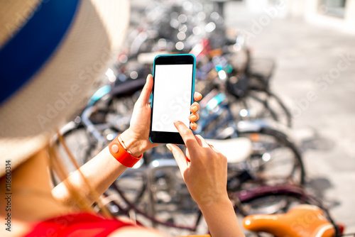 Woman holding smart phone with white screen on the bicycle parking background. Bicycle rent with smart phone. Mobile travel program