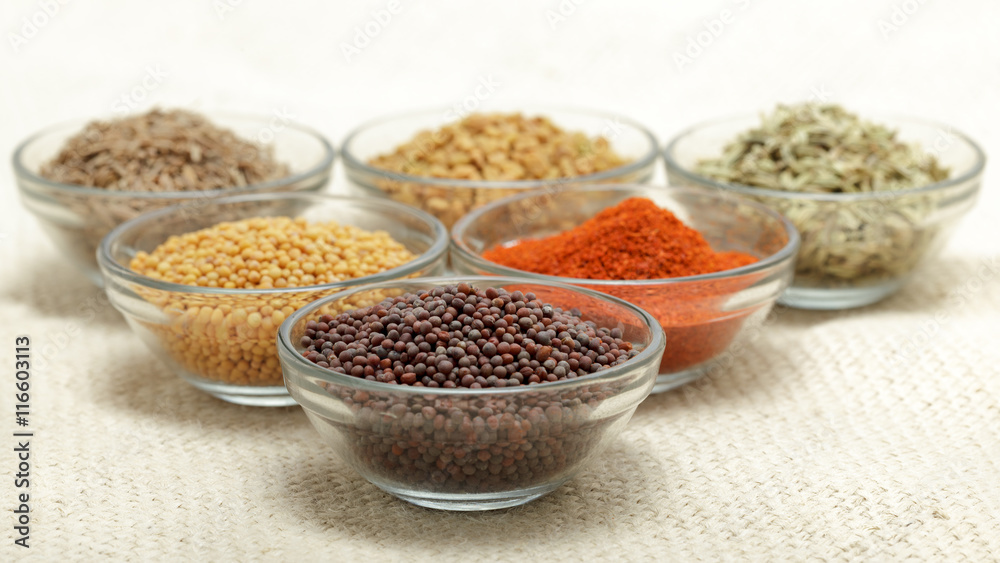 Different types of Indian spices in glass bowl, focus on brown mustard seeds on jute mat background. Front view.