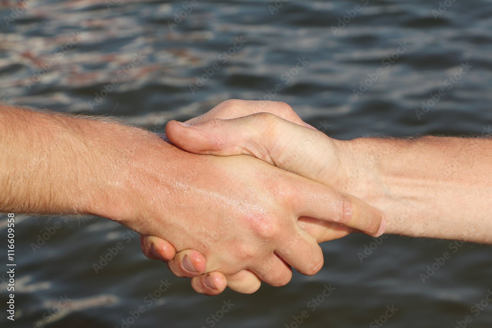 Suntanned male hands make handshake against a water surface Suntanned male hands make handshake against a water surface