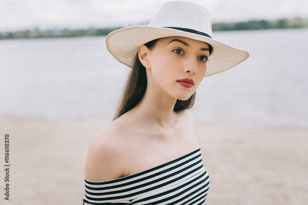 Stylish and fashionable girl model in white hat posing on the beach in summer.