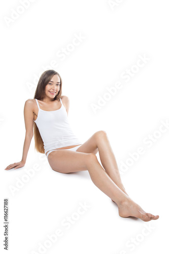 Beautiful attractive Caucasian young model with long legs sitting against white background and looking at camera. Cheerful smiling person posing in tank top and panties. Vertical studio portrait shot © fizkes