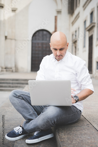 Young handsome caucasian businessman sitting on a staircase using a laptop leaning on his knees, looking downward the screen, pensive - working, thoughtful, busy concept