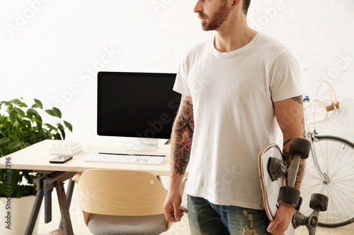 Brutal tattoooed man in blank t-shirt holds surf skateboard in big loft in front of his working desktop with computer Vintage bicycle parked on background.