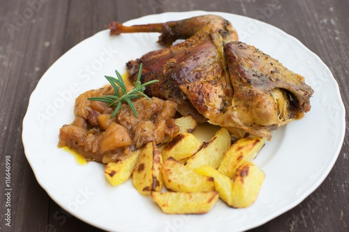 Roasted or baked Guinea fowl served with baked potatoes and sweet onion with apples and raisins. Meal spiced by rosemary and black pepper is served on the white plate and old rustic wooden table.