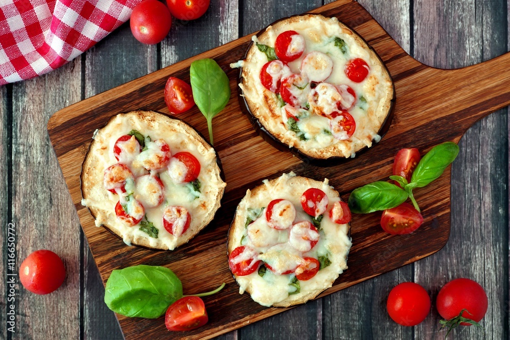 Healthy eggplant mini pizzas with melted mozzarella, tomatoes and basil on a serving board against rustic wood