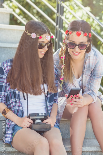 Portrait of two young girlfriends on summer vacation using smart phone and vintage camera. They are sitting on the outdoors stairs and smiling to camera. 