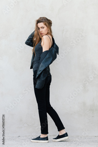 Happy young beautiful woman in black leather jacket black jeans slip-on posing for model tests against textured wall