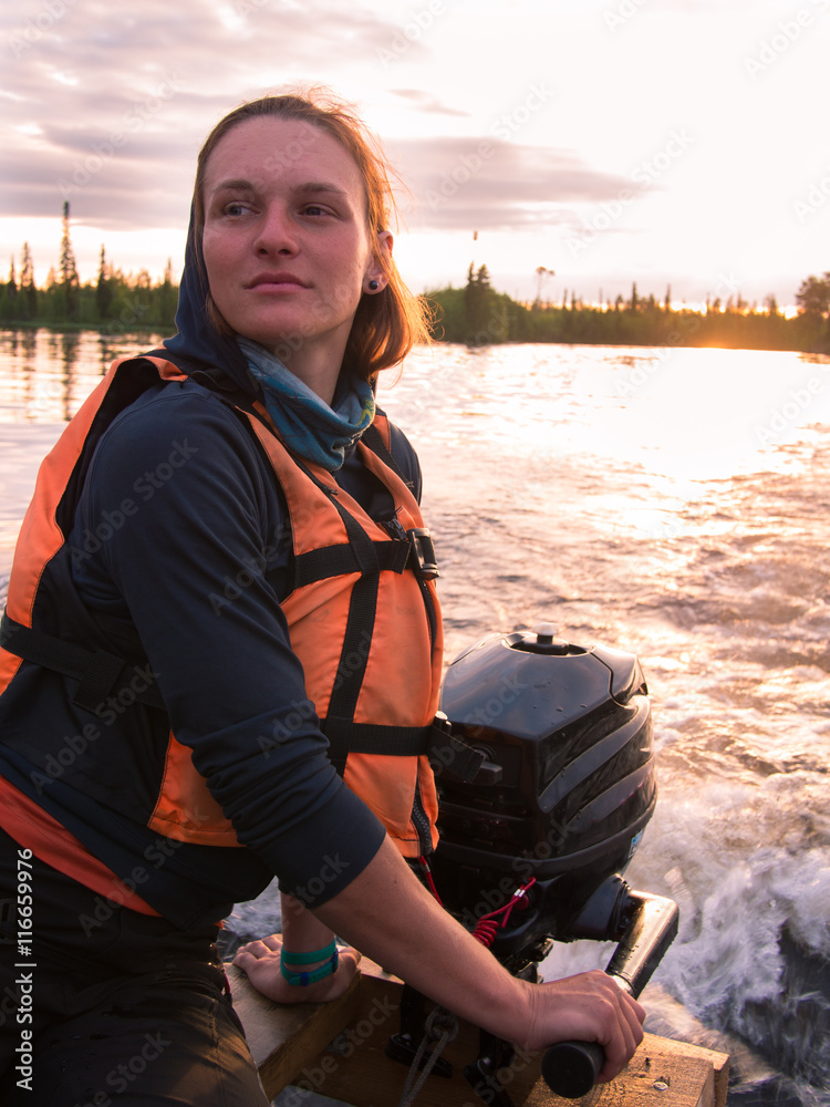 Fototapeta premium Young girl in life jacket steers the boat with a motor at sunset with clouds