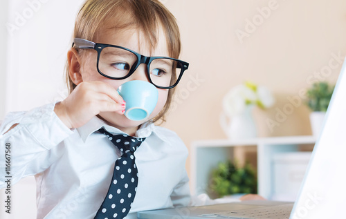 Photography Smart toddler girl with glasses drinking coffee while using a laptop