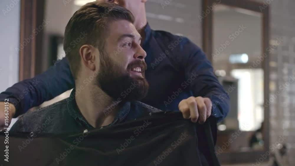 Barber smiling and talking to his client while covering him with cloth to prepare for haircut