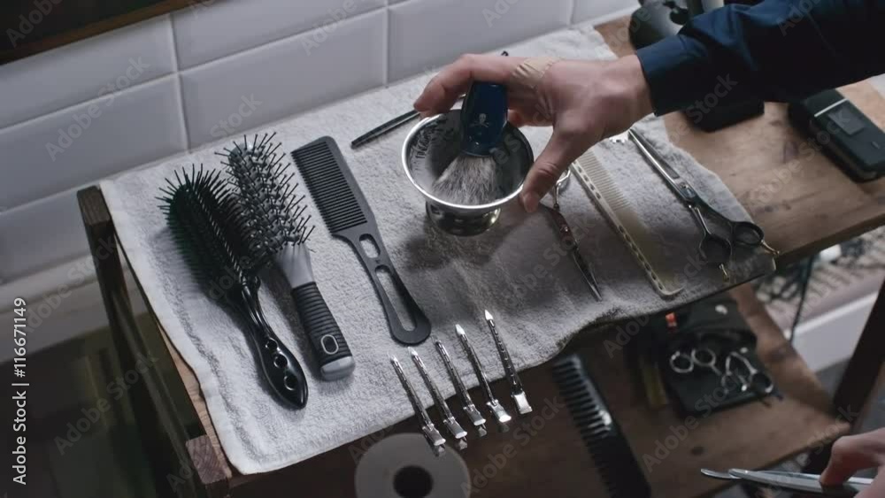 High angle view of barbershop tools on the shelve and male hands taking straight razor and placing bowl with shaving cream and brush in it